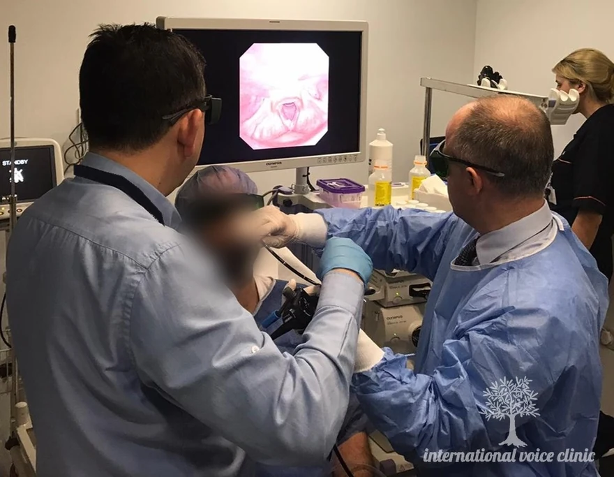 A seated, awake patient undergoing laser treatment through a flexible endoscope passed through the nose, with the laryngeal view shown on a monitor behind and everyone in the room wearing laser safety glasses.
